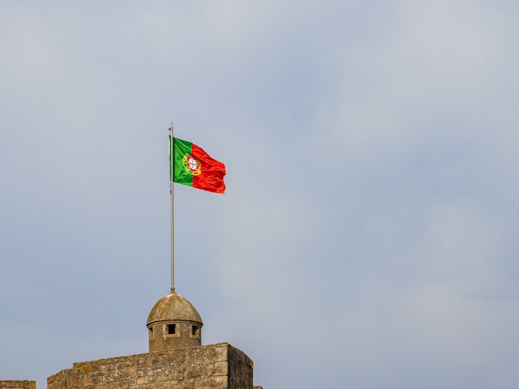 A Portuguese flag is flying atop a stone structure with a dome-shaped lookout. The sky in the background is overcast with light clouds.