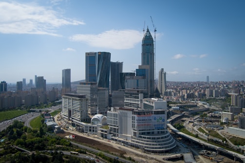 A sprawling urban landscape featuring a cluster of modern skyscrapers with varied architectural styles. The tallest building is under construction, with cranes visible at the top. Surrounding the high-rise structures are smaller buildings, roads, and green spaces. The sky is partly cloudy, creating a bright and open atmosphere.