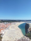 An expansive beach stretches along the coastline, filled with rows of sun umbrellas and crowds of people. The sandy beach is bordered by a town with white buildings and terracotta rooftops. A clear blue sky looms overhead, and a large rock formation is visible in the foreground.