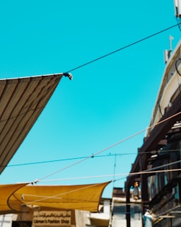 An urban scene with several awnings and wires stretching across the frame against a clear blue sky. Below, there are storefronts and buildings with a focus on a shop named 'Usman's Fashion Shop' in the background.