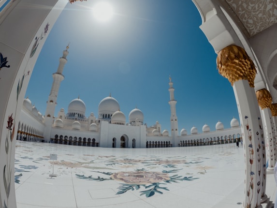 A grand, ornate courtyard of a mosque with a clear blue sky and bright sunlight. The architecture features large white domes, tall minarets, and intricately designed columns with floral patterns. The floor is decorated with artistic floral designs.