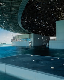 An architectural structure features a large, intricate roof patterned with geometric openings. The building sits beside what appears to be a body of water, reflecting the blue sky overhead. The roof casts shadows and dappled light across the surfaces. In the distance, construction cranes and modern buildings outline the sky.