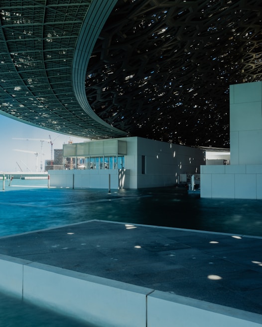 An architectural structure features a large, intricate roof patterned with geometric openings. The building sits beside what appears to be a body of water, reflecting the blue sky overhead. The roof casts shadows and dappled light across the surfaces. In the distance, construction cranes and modern buildings outline the sky.