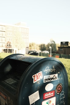 A blue postal mailbox is in the foreground with various stickers and graffiti on it. Behind the mailbox, there is a grassy area and a few trees with a multi-story building in the background under a clear sky.