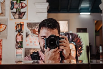 A person is taking a photo with a DSLR camera, capturing their reflection in a mirror. The background features a variety of colorful paintings and artwork, creating an artistic and vibrant atmosphere.