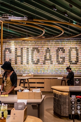 A modern cafe interior with a large wall mural that spells out 'CHICAGO' using various small tiles. The ceiling features a series of green beams and circular lighting fixtures. A barista in an apron stands at the counter, while another person is using a device at a nearby table.