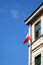 A corner of a building with a Polish flag attached to a pole extending from the facade under a clear blue sky. The building has beige walls with green window frames and decorative architectural elements.