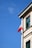 A corner of a building with a Polish flag attached to a pole extending from the facade under a clear blue sky. The building has beige walls with green window frames and decorative architectural elements.