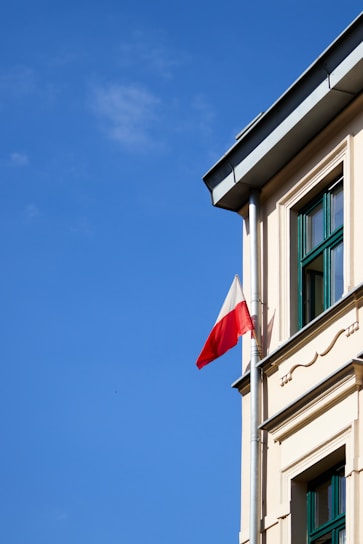 A corner of a building with a Polish flag attached to a pole extending from the facade under a clear blue sky. The building has beige walls with green window frames and decorative architectural elements.
