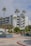 A modern multi-story apartment building with balconies surrounded by tall palm trees under a partly cloudy sky. The courtyard features landscaped planters with small trees and shrubs, and the road is lined with red and blue curbs.