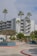 A modern multi-story apartment building with balconies surrounded by tall palm trees under a partly cloudy sky. The courtyard features landscaped planters with small trees and shrubs, and the road is lined with red and blue curbs.
