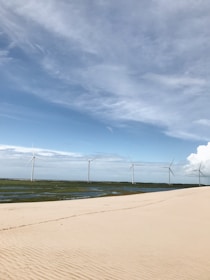 A serene landscape featuring a sandy foreground leading to a row of wind turbines on lush green terrain under a vast sky with scattered clouds.