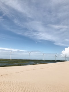 A serene landscape featuring a sandy foreground leading to a row of wind turbines on lush green terrain under a vast sky with scattered clouds.