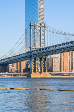 A large suspension bridge spans across a river, with a tall modern skyscraper and several residential buildings visible in the background. The bridge's intricate steel framework is prominently featured, and the water reflects the blue of the clear sky.