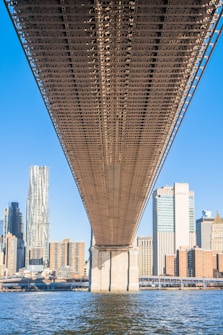 A large steel bridge spans across a body of water, with the bridge's intricate metal framework prominently showcased. In the background, a city skyline is visible, featuring modern skyscrapers and a mix of architectural styles. The sky is clear and blue, indicating a sunny day.
