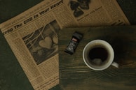 A rustic wooden table holds a cup of coffee, a newspaper, and a wrapped bar of dark chocolate. The newspaper features a large headline with inspirational content and an image of hanging heart decorations. The cup of coffee is dark, with a light froth on top, positioned next to the chocolate bar that displays '55% dark' on its wrapper.