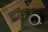 A rustic wooden table holds a cup of coffee, a newspaper, and a wrapped bar of dark chocolate. The newspaper features a large headline with inspirational content and an image of hanging heart decorations. The cup of coffee is dark, with a light froth on top, positioned next to the chocolate bar that displays '55% dark' on its wrapper.
