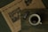 A rustic wooden table holds a cup of coffee, a newspaper, and a wrapped bar of dark chocolate. The newspaper features a large headline with inspirational content and an image of hanging heart decorations. The cup of coffee is dark, with a light froth on top, positioned next to the chocolate bar that displays '55% dark' on its wrapper.