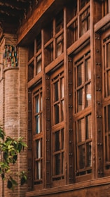 A traditional wooden-framed window with intricate detailing set against a backdrop of a brick wall. The window features multiple glass panes and decorative metallic knobs. Near the lower left corner, a small green tree adds a touch of nature.