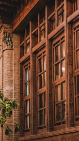 A traditional wooden-framed window with intricate detailing set against a backdrop of a brick wall. The window features multiple glass panes and decorative metallic knobs. Near the lower left corner, a small green tree adds a touch of nature.