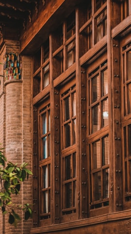 A traditional wooden-framed window with intricate detailing set against a backdrop of a brick wall. The window features multiple glass panes and decorative metallic knobs. Near the lower left corner, a small green tree adds a touch of nature.