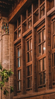 A traditional wooden-framed window with intricate detailing set against a backdrop of a brick wall. The window features multiple glass panes and decorative metallic knobs. Near the lower left corner, a small green tree adds a touch of nature.