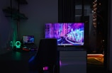 A dimly lit room with a dark theme, featuring a desk setup that includes a laptop and a large monitor displaying a vibrant cityscape at night. The neon colors on the screens provide a cyberpunk aesthetic. A gaming headset with glowing accents is placed beside the laptop. Shelves with small items are mounted on the wall, adding to the tech ambiance.