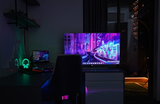 A dimly lit room with a dark theme, featuring a desk setup that includes a laptop and a large monitor displaying a vibrant cityscape at night. The neon colors on the screens provide a cyberpunk aesthetic. A gaming headset with glowing accents is placed beside the laptop. Shelves with small items are mounted on the wall, adding to the tech ambiance.