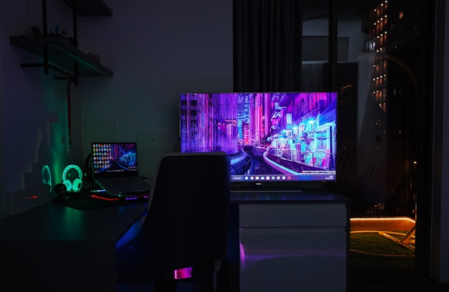 A dimly lit room with a dark theme, featuring a desk setup that includes a laptop and a large monitor displaying a vibrant cityscape at night. The neon colors on the screens provide a cyberpunk aesthetic. A gaming headset with glowing accents is placed beside the laptop. Shelves with small items are mounted on the wall, adding to the tech ambiance.