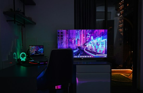 A dimly lit room with a dark theme, featuring a desk setup that includes a laptop and a large monitor displaying a vibrant cityscape at night. The neon colors on the screens provide a cyberpunk aesthetic. A gaming headset with glowing accents is placed beside the laptop. Shelves with small items are mounted on the wall, adding to the tech ambiance.