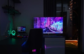 A dimly lit room with a dark theme, featuring a desk setup that includes a laptop and a large monitor displaying a vibrant cityscape at night. The neon colors on the screens provide a cyberpunk aesthetic. A gaming headset with glowing accents is placed beside the laptop. Shelves with small items are mounted on the wall, adding to the tech ambiance.