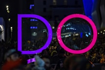 Large illuminated letters spelling 'DO' stand prominently against a nighttime urban backdrop, surrounded by a crowd of people. The letter 'D' is illuminated in purple, while the 'O' is bright pink. City lights and reflections add to the vibrant atmosphere, with the crowd appearing lively and engaged.