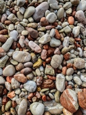 A collection of smooth, rounded pebbles in various sizes, colors, and textures. The stones range from earthy reds and browns to soft greys and off-whites, with some displaying intricate patterns and natural markings.