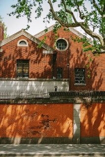 A brick building with distinct architectural features, including arched windows and sharp rooflines. Shadows of tree branches are cast on the bricks, creating a patterned effect. In the foreground, a brick wall topped with metal spikes runs parallel to the building.