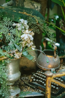 An old typewriter surrounded by lush, green vegetation and a variety of white and wilted flowers in a metallic vase, with a vintage kettle nearby. The scene gives a sense of being in an overgrown garden or a forgotten corner filled with antique objects.
