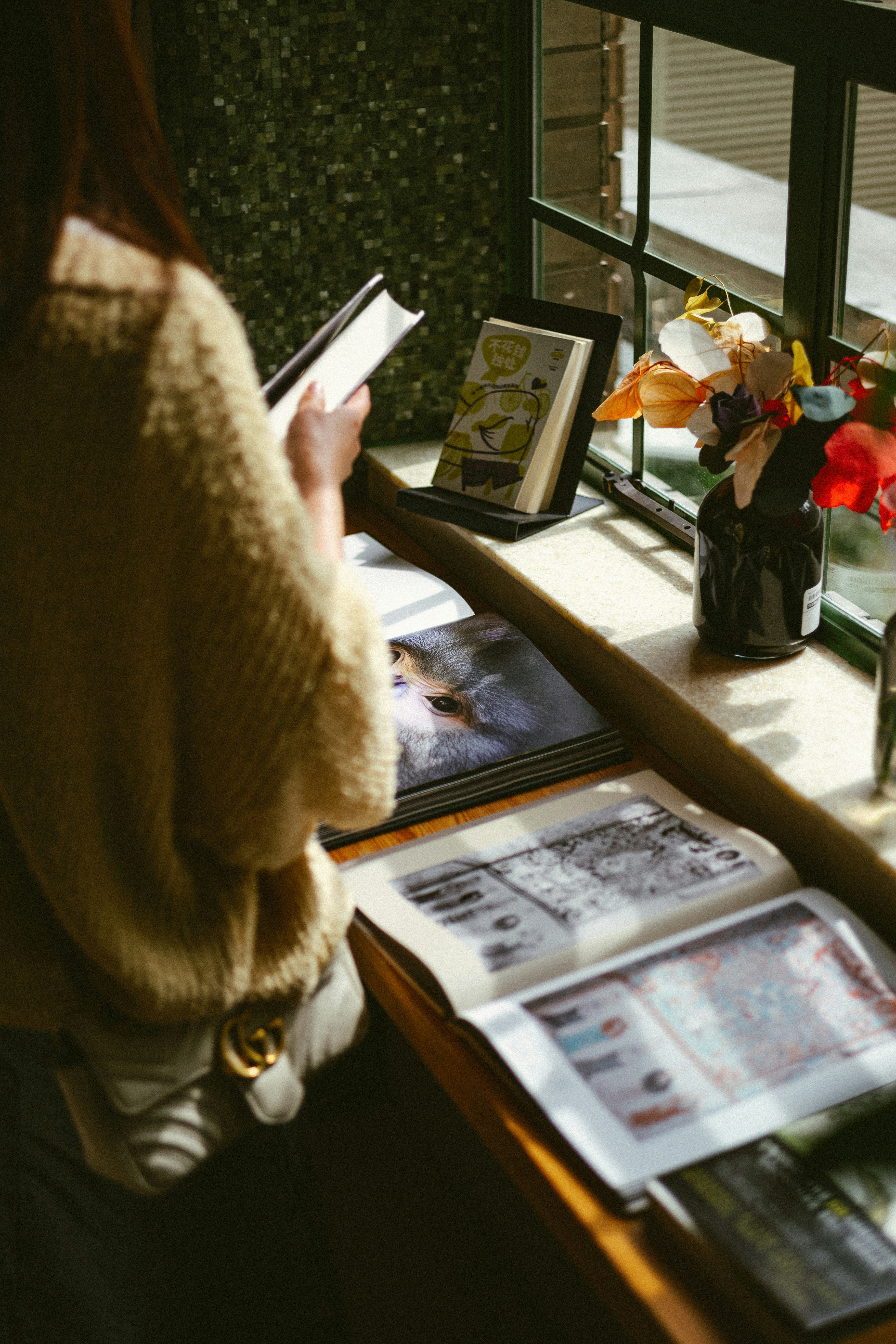A person reads a book by a window with flowers.