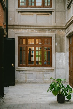 The image presents a courtyard scene featuring a plant in a pot placed on a tiled floor. A building facade with large wooden framed windows occupies the background, displaying variations of grey and brown tones. The mix of natural and architectural elements creates a serene and structured atmosphere.
