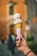 A hand holds a cone with two scoops of ice cream, speckled with small pieces. The cone is wrapped in branded paper with 'Dal Cuore' printed on it. Two green sticks are inserted into the ice cream. A blurred background features a warm-toned building.