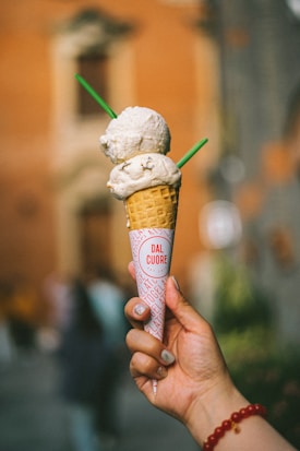 A hand holds a cone with two scoops of ice cream, speckled with small pieces. The cone is wrapped in branded paper with 'Dal Cuore' printed on it. Two green sticks are inserted into the ice cream. A blurred background features a warm-toned building.