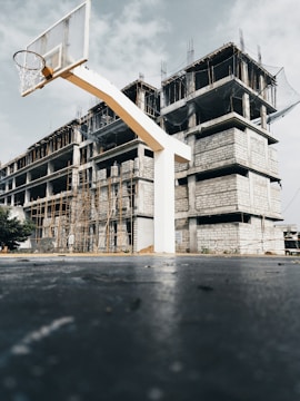 A basketball hoop stands in the foreground on a rough court, with a partially constructed building in the background. The building is made of concrete blocks and is surrounded by scaffolding. The sky is overcast, adding a muted tone to the scene.