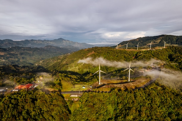Wind turbines are positioned across a lush, hilly landscape with dense green vegetation. Low-lying clouds create a misty effect, and a few small buildings with red rooftops are scattered among the hills. In the background, mountain ranges stretch into the distance under a partly cloudy sky.