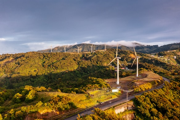 An expansive landscape of lush green hills topped with a series of tall wind turbines. The turbines are aligned along a ridgeline, catching the light of a setting or rising sun. Clouds gently rest on the hills in the background, adding depth and texture to the scene.