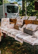 A display of art prints and notebooks is arranged on a table, with various landscape-oriented prints placed in wooden crates and baskets. The tablecloths also feature colorful prints, and a white van is parked in the background, partially obscured by trees.