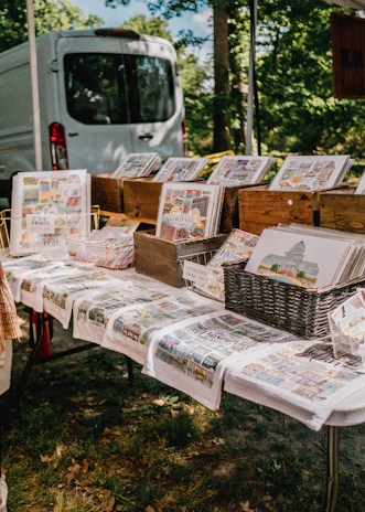 A display of art prints and notebooks is arranged on a table, with various landscape-oriented prints placed in wooden crates and baskets. The tablecloths also feature colorful prints, and a white van is parked in the background, partially obscured by trees.