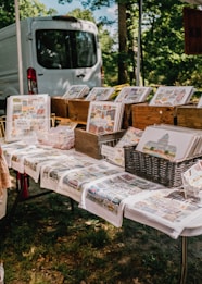 A display of art prints and notebooks is arranged on a table, with various landscape-oriented prints placed in wooden crates and baskets. The tablecloths also feature colorful prints, and a white van is parked in the background, partially obscured by trees.
