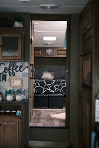 A cozy interior of a camper features wooden cabinetry and a small counter with a coffee setup. Through a doorway, a bed is visible with a black and white patterned bedspread and pillows, along with storage compartments underneath. Soft lighting from the ceiling complements the warm and inviting atmosphere.