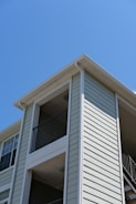 The exterior of a light-colored, multi-story residential building with siding and balconies features prominently. The architecture includes clean lines and a modern design, with visible railings on the upper floors. The building is set against a clear blue sky, suggesting a sunny day.