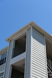 The exterior of a light-colored, multi-story residential building with siding and balconies features prominently. The architecture includes clean lines and a modern design, with visible railings on the upper floors. The building is set against a clear blue sky, suggesting a sunny day.