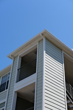 The exterior of a light-colored, multi-story residential building with siding and balconies features prominently. The architecture includes clean lines and a modern design, with visible railings on the upper floors. The building is set against a clear blue sky, suggesting a sunny day.