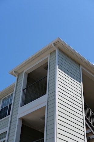 The exterior of a light-colored, multi-story residential building with siding and balconies features prominently. The architecture includes clean lines and a modern design, with visible railings on the upper floors. The building is set against a clear blue sky, suggesting a sunny day.